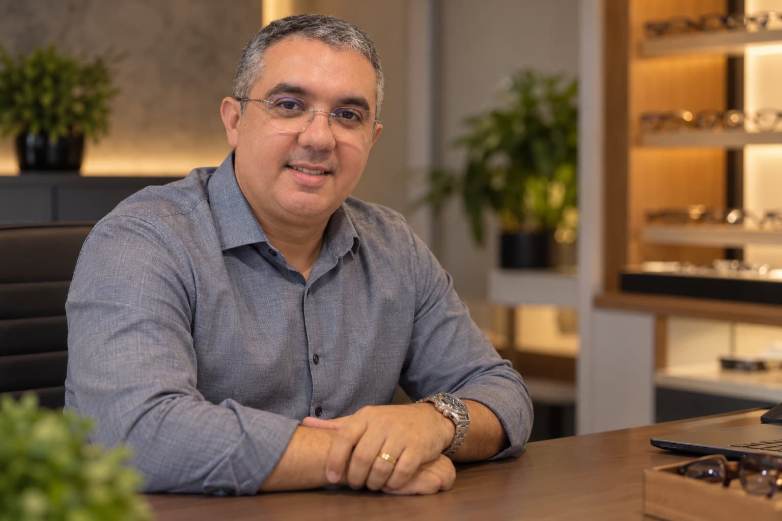 Tiago Souza, consultor óptico, sentado à mesa de atendimento, com prateleiras de armações iluminadas ao fundo